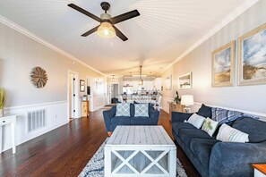 Ein geräumiges Wohnzimmer mit Holzböden, Deckenventilator, einem weißen Couchtisch, einem dunkelblauen Sofa und einer Küche im Hintergrund.