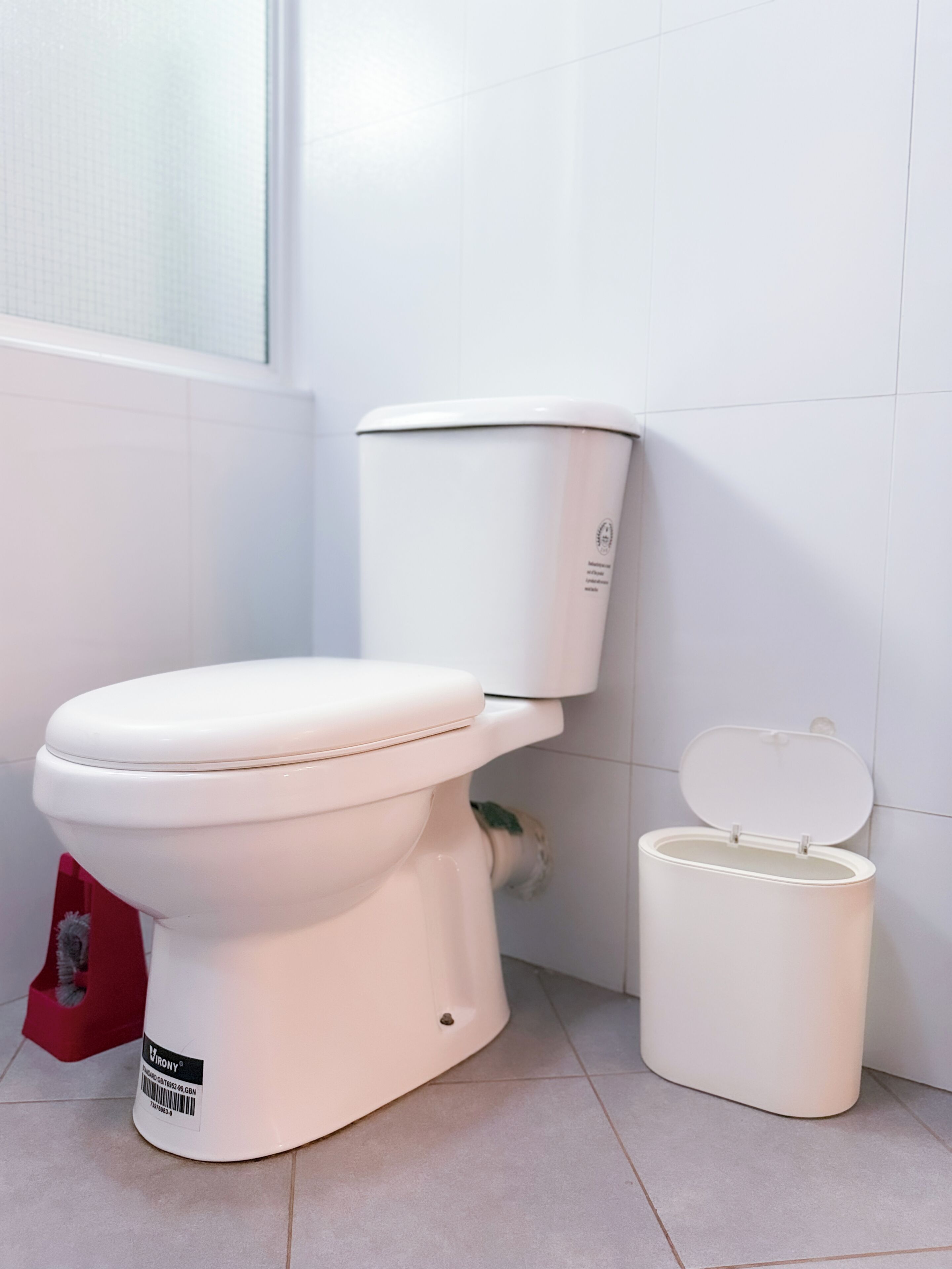 A white toilet with a closed lid, a white toilet tank, and a white trash bin in a bathroom with white tiled walls and floor.