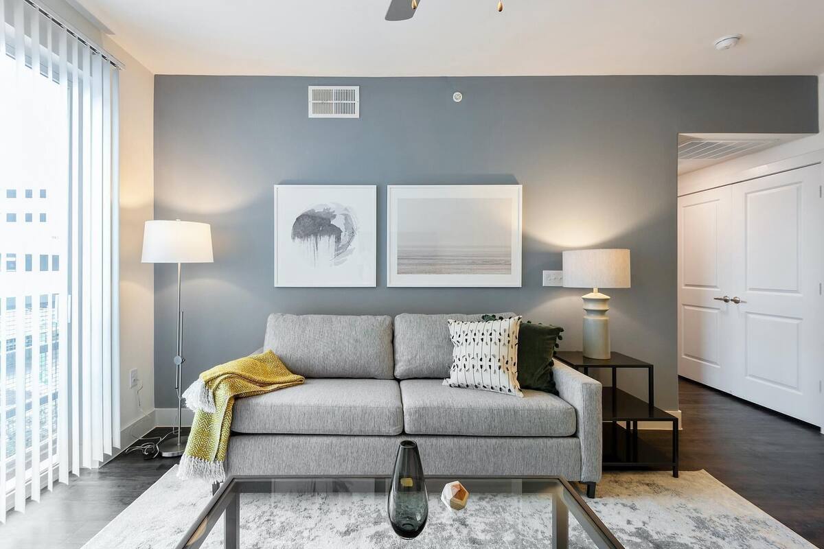A modern living room with a grey sofa, two framed artworks, a floor lamp, and a glass coffee table.