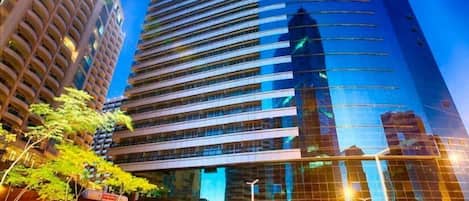 A modern high-rise building with a reflective glass facade, surrounded by greenery and other buildings.
