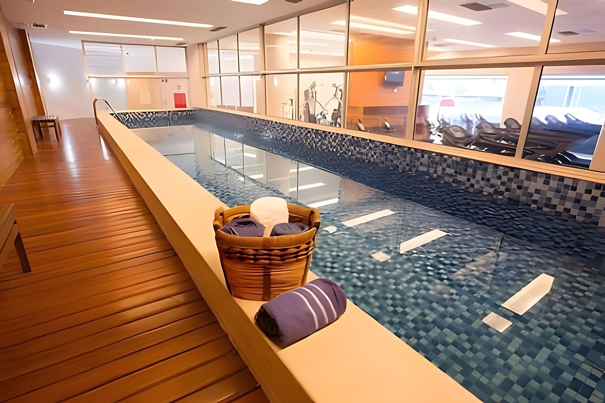 Piscina interior moderna com paredes de vidro transparente, piso de madeira e área de estar com uma cesta de toalhas.