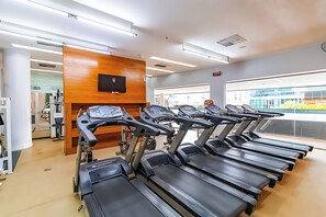 Una palestra con diverse tapis roulant disposti in file, una reception in legno e un'ampia finestra con vista sugli edifici esterni.