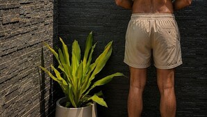 Une personne se tient dans un espace douche, avec un mur en briques noires, une plante en pot et un tapis bleu sur le sol.