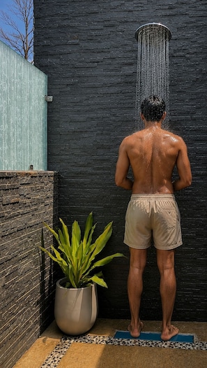 A person standing in a shower area with a black brick wall, a potted plant, and a blue mat on the floor. - Kayo Resort (Kurai)
