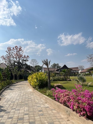 Uma trilha pavimentada leva a um edifício com telhado de azulejos, rodeado por vegetação e plantas floridas.