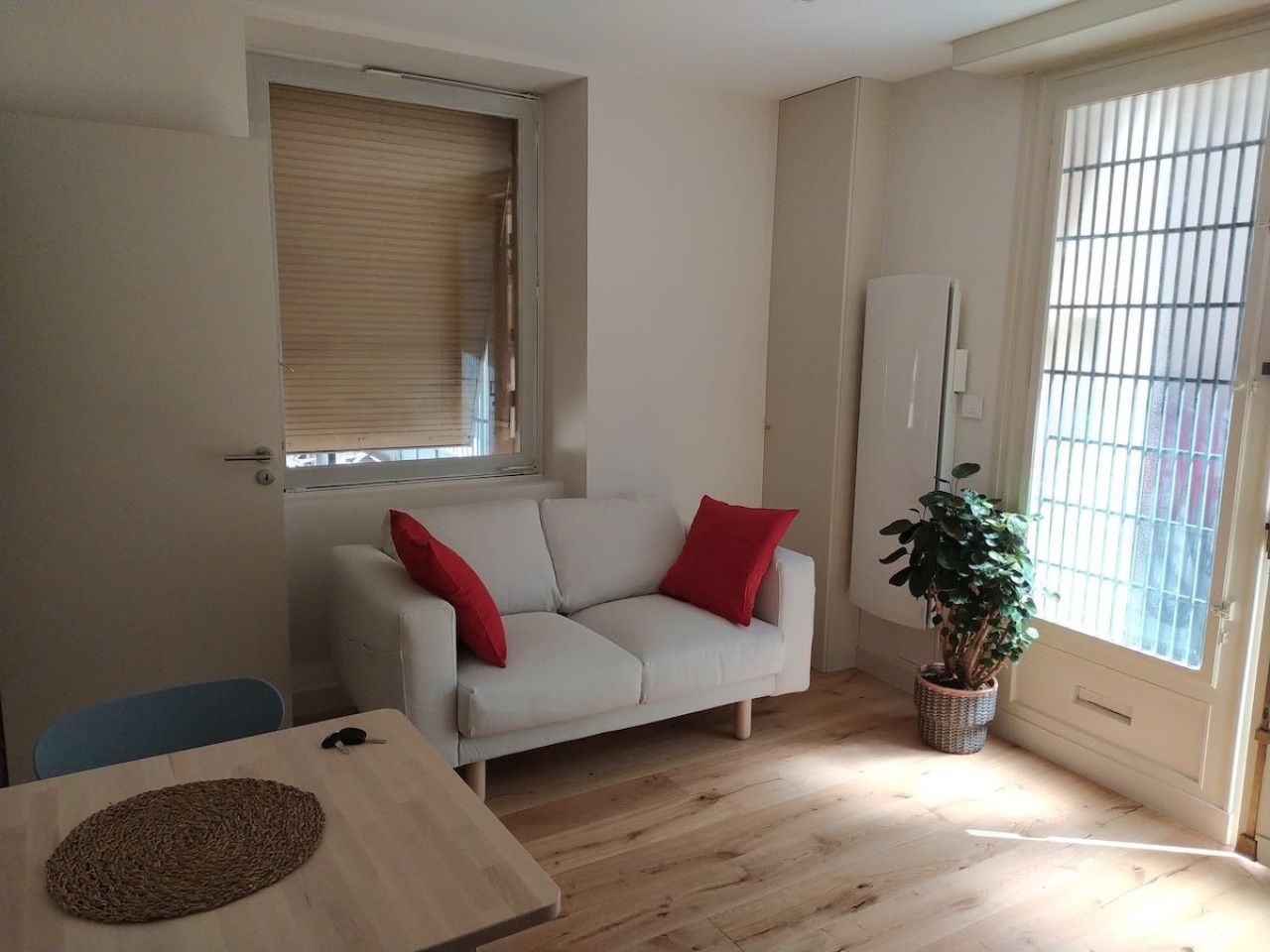 Una habitación con un sofá blanco, cojines rojos, una mesita, una planta en maceta y una ventana con persianas.