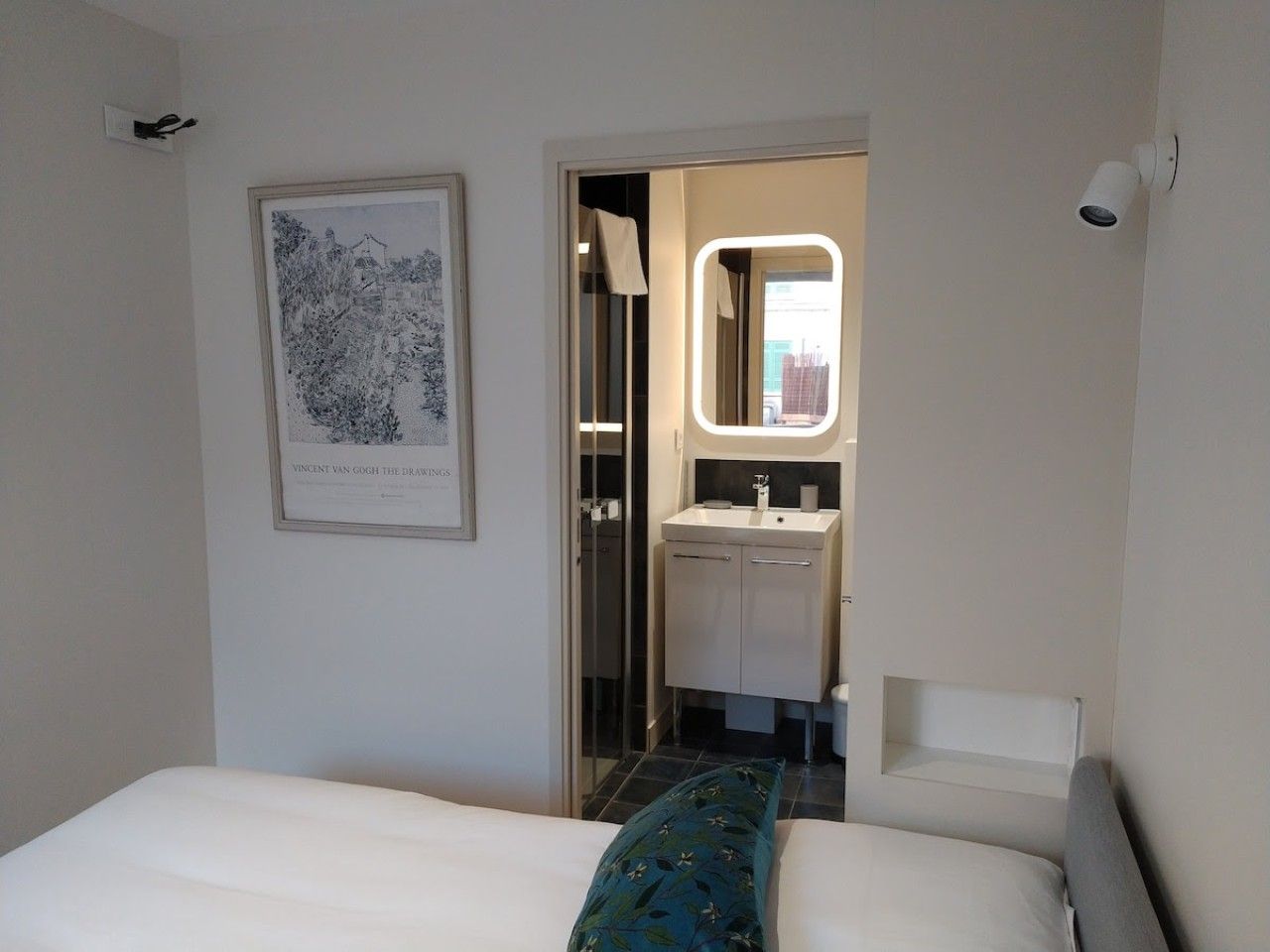 A hotel room with a bed, a framed artwork, a bathroom with a mirror, and a white wall.