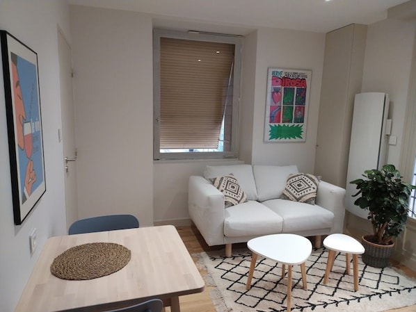 A living room with a sofa, two white side tables, a wooden coffee table, a patterned rug, a potted plant, and a window with blinds.