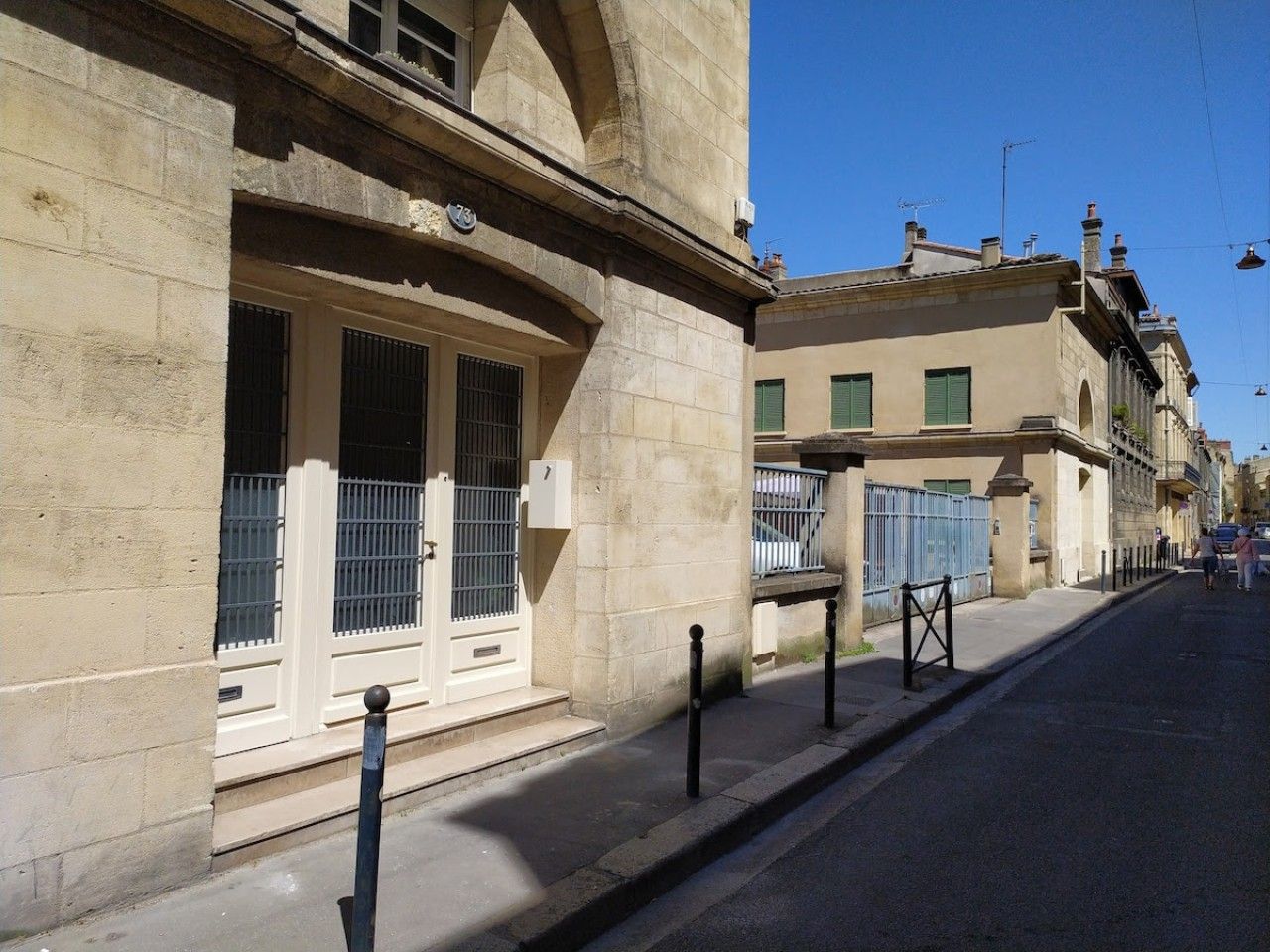 A street view with a building featuring a large window with a metal grille, a balcony, and a door with a small mailbox.