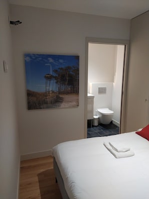 A bedroom with a bed, a painting of a landscape, a bathroom with a toilet and sink, and a wooden floor.