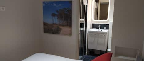 Une chambre d’hôtel avec un lit, un oreiller rouge, une serviette blanche, un tableau et une salle de bain visible à travers une porte ouverte.