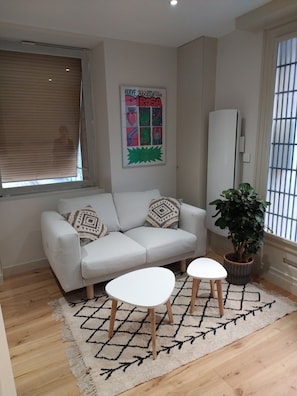 A modern living room with a white sofa, two white side tables, a patterned rug, a potted plant, and a colorful poster on the wall.