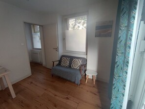 A room with a wooden floor, a sofa with patterned cushions, a small round table, and a window with a view of trees. - One bedroom  in France, Bordeaux (Bordeaux)