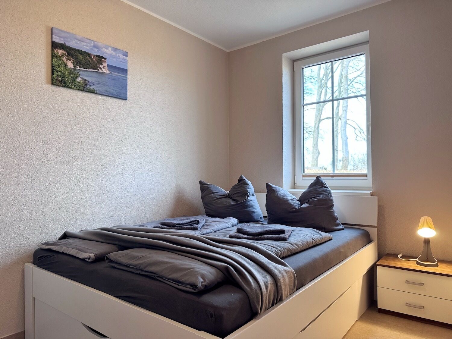 A bedroom with a bed, pillows, a nightstand, a lamp, a window, and a painting on the wall.