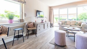 Ein modernes Wohnzimmer mit einer Couch, einem Couchtisch, Sesseln, einem Fernseher und einem großen Fenster mit Blick auf Grünflächen.