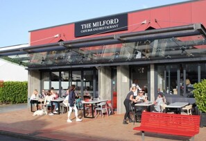 Ein Außenbereich des 'Milford Cafe Bar and Restaurant' mit Tischen, Stühlen und einer roten Bank, außerdem ein Hund.