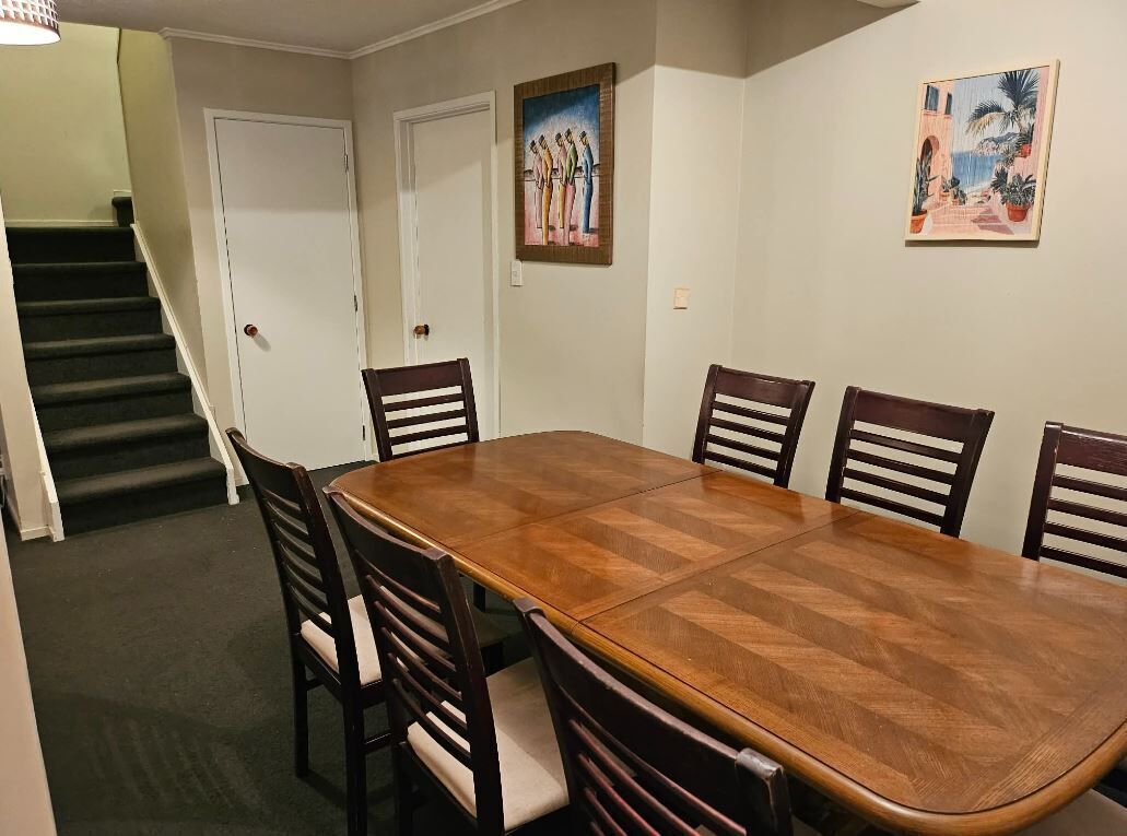 Une salle à manger avec une grande table en bois et six chaises, un escalier et deux tableaux accrochés aux murs.