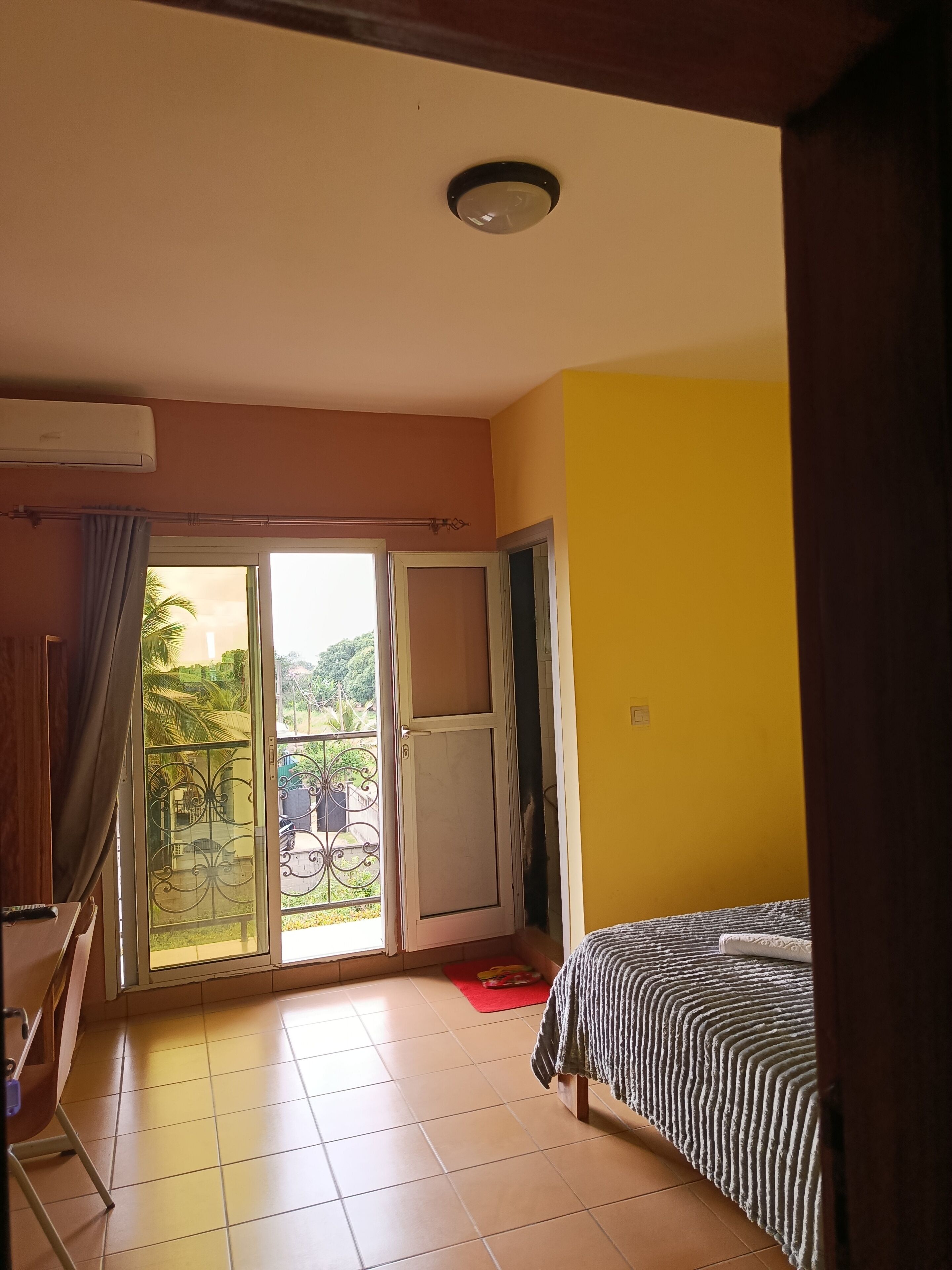Une chambre dotée d’un lit, d’une porte-fenêtre coulissante donnant sur un balcon, d’un ventilateur de plafond et d’un sol carrelé.