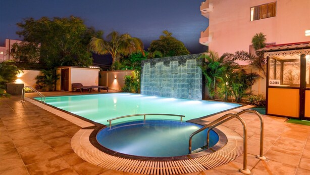 Una piscina con pared de azulejos y un jacuzzi, rodeada de palmeras y un edificio con cartel de cerrado.