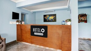 Hotellin vastaanottotiski, jossa on kyltti, jossa lukee ”HOTELO Company-Serviced”, sekä taulutelevisio, maailmankartta ja pieni ruukkukasvi.