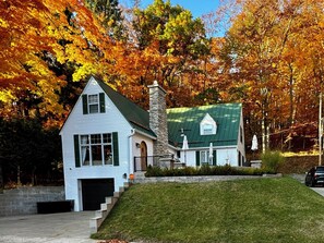 Una casa de dos pisos con techo verde y chimenea de piedra, rodeada de follaje otoñal.