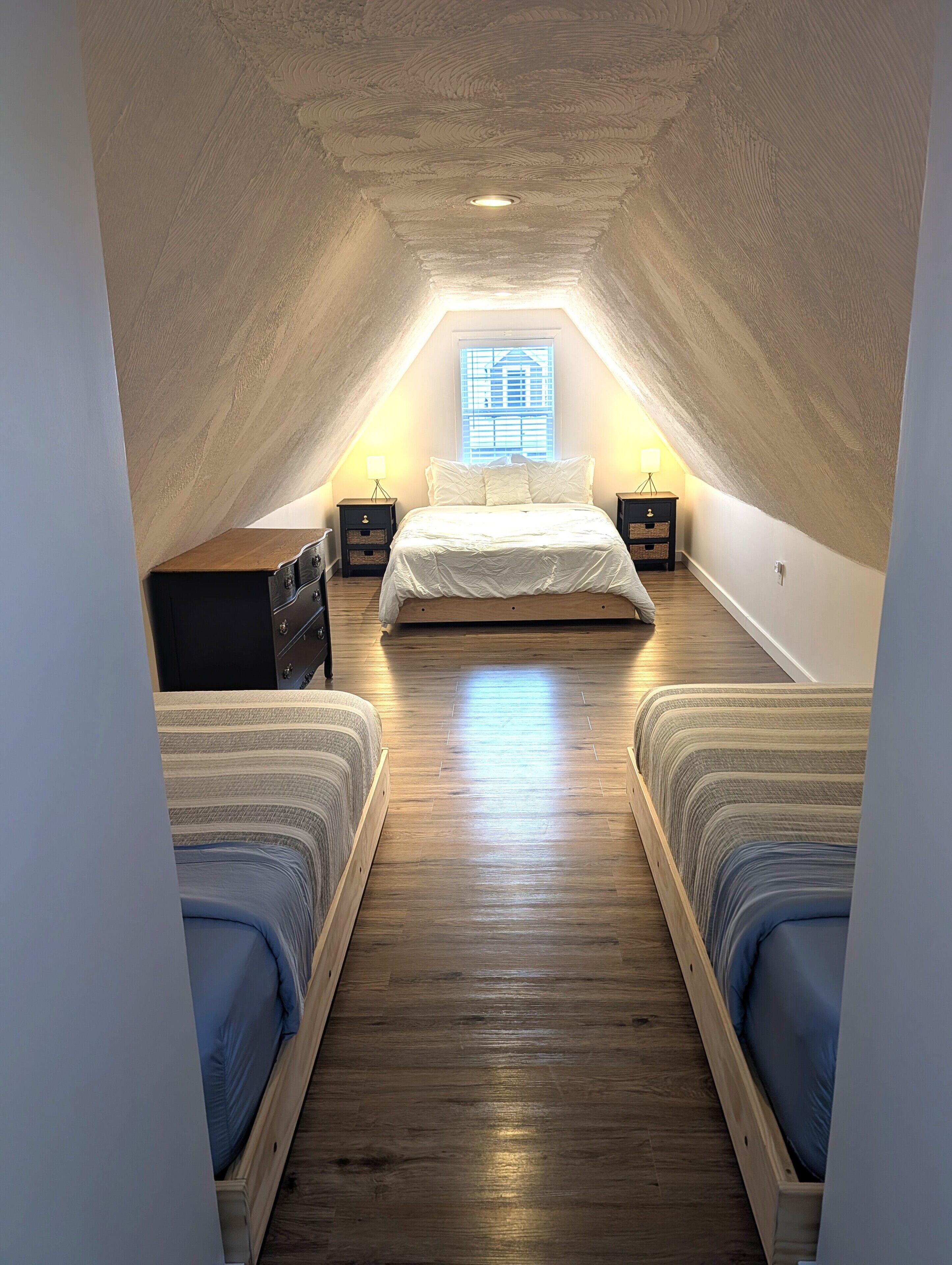 Une petite chambre mansardée, chaleureuse et accueillante, avec un plafond en pente, un lit placé au centre, deux tables de chevet et une fenêtre équipée de stores.