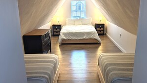 Un dormitorio acogedor en el ático, con techo inclinado, una cama central, dos mesitas de noche y una ventana con persianas.