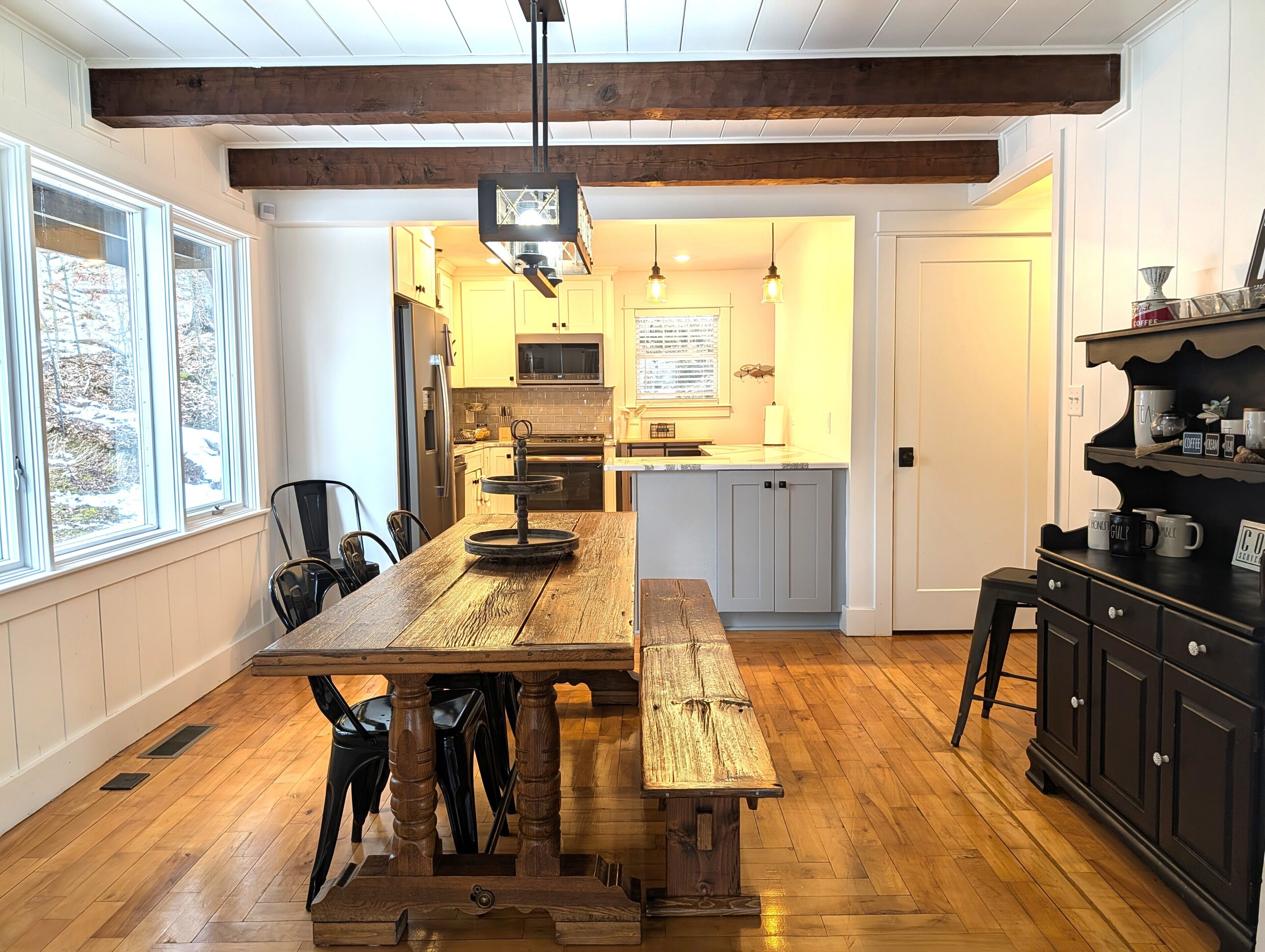 A kitchen with wooden beams, a large wooden table, and a black cabinet with mugs.