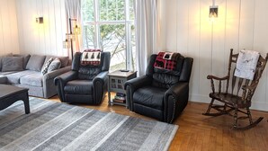 Una sala de estar acogedora con un sofá, dos sillones de cuero, un mecedora de madera, un tapete estampado y una ventana con cortinas.