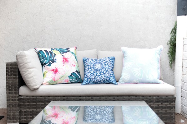 A sofa with three pillows, a glass table, and a brick wall.