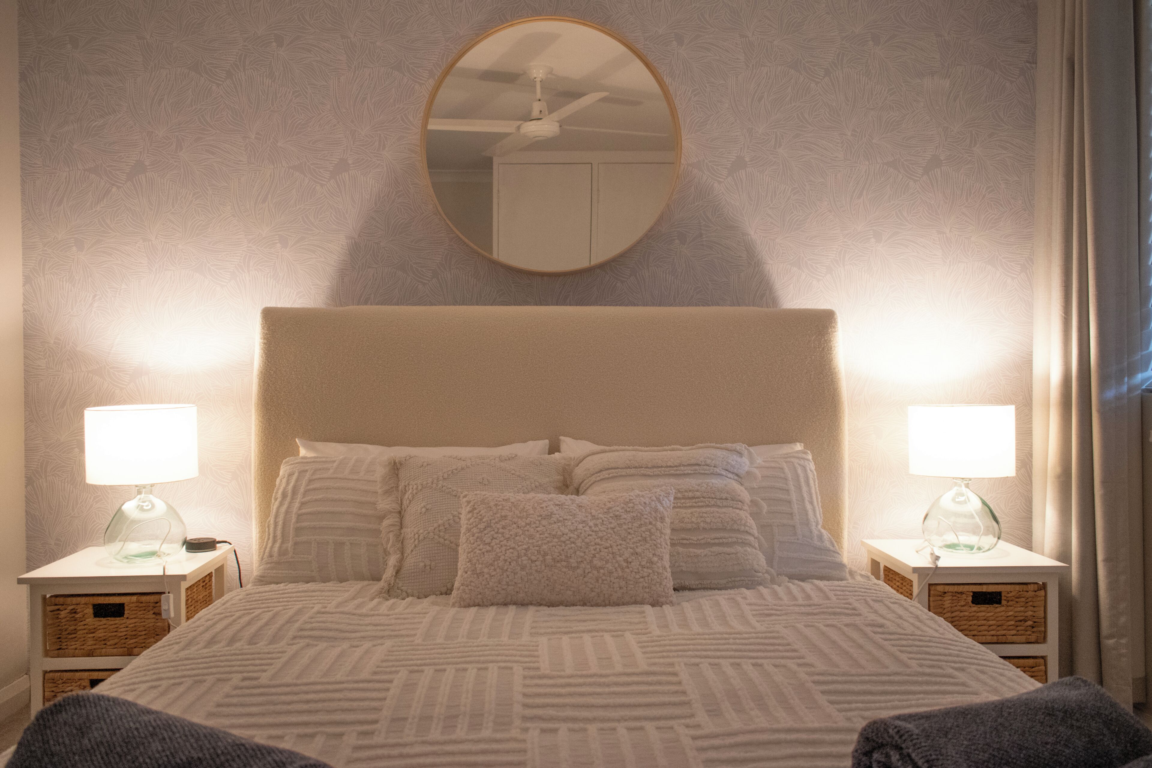 A neatly made bed with a patterned bedspread, two bedside lamps, a round mirror, and a ceiling fan.