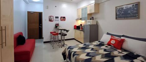 A compact hotel room with a bed, a red sofa, a kitchenette, and a small dining area.
