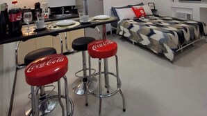 Ein kleines Zimmer mit Küchenzeile, einem Bett mit gemustertem Überwurf und zwei Barhocker mit roten Sitzflächen.