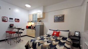 Ein kompaktes Hotelzimmer mit Bett, einer kleinen Küchenzeile, einer Essecke mit Hocker und einem Nachttisch.