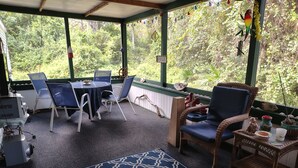 Ein überdachter Außenbereich mit Tisch und Stühlen, ein Korbstuhl, ein kleiner Tisch mit einer Dekoration sowie ein Vogelhaus.