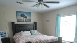 Ein Schlafzimmer mit einem Bett, einem Deckenventilator, einem Fenster mit Vorhängen und einem Bild an der Wand.