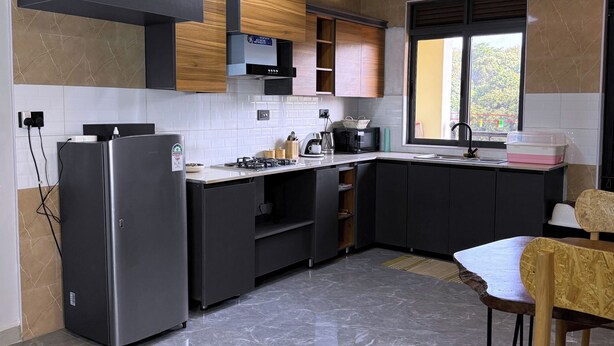 A modern kitchen with a built-in refrigerator, dark cabinetry, a window with a view, and a dining area with a chair. - Constatia Homes (Kampala)