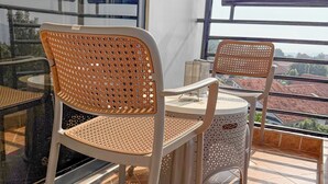 A balcony with two wicker chairs and a small table, a tiled floor, and a view of buildings through large windows. - Constatia Homes (Kampala)