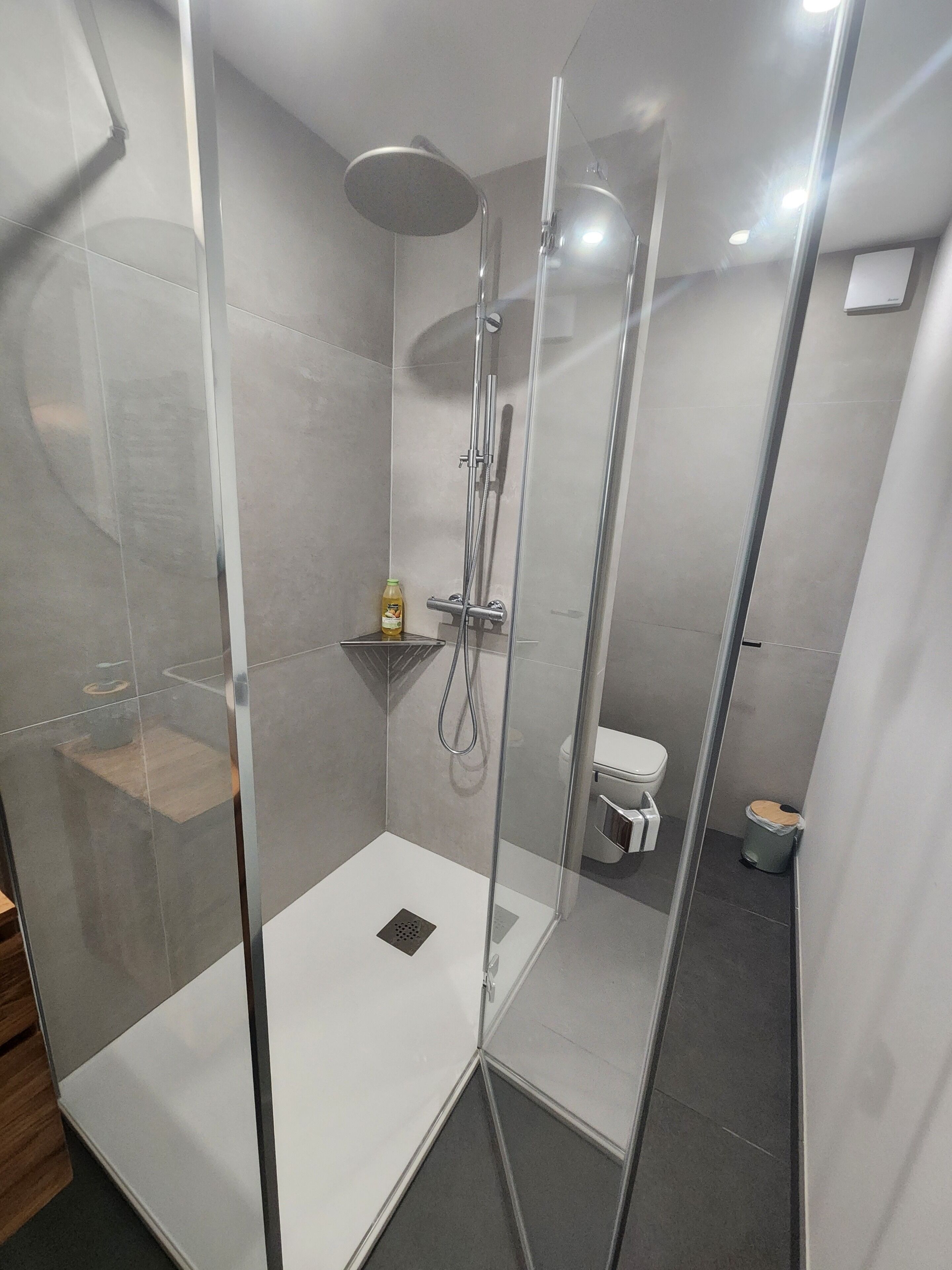 Moderni kylpyhuone, jossa on lasiseinällä varustettu suihku, wc ja pieni hylly, jossa on hygieniatuotteita.