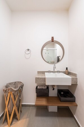 Une salle de bain avec un lavabo blanc, un miroir rond, un porte-serviettes, une poubelle à rayures et une étagère en bois.