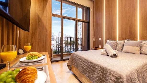 Habitación de hotel con una cama grande, una mesa de desayuno con croissants y fruta, y vistas a la ciudad a través de amplios ventanales.