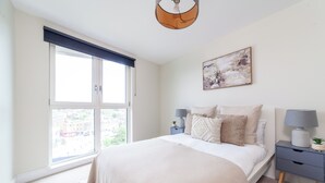 Un dormitorio con un ventanal grande, una cama con cubrecama blanco, una mesita de noche con lámpara y un cuadro abstracto colgado en la pared.