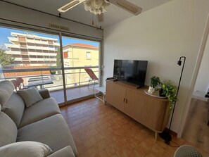 Een zithoek met een bank, een tv op een houten kast, een kamerplant en een uitzicht op een gebouw door grote ramen.