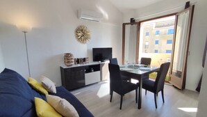 Een moderne woonkamer met een bank, een eettafel met stoelen, een televisie en een balkon met uitzicht op gebouwen.