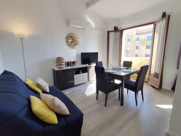 Ein modernes Wohnzimmer mit einer Couch, einem Esstisch mit Stühlen, einem Fernseher und einem Balkon mit Blick auf Gebäude.