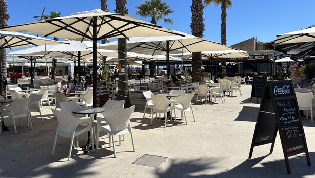 Een terras met witte plastic stoelen en parasols, palmbomen en een Coca-Cola bord.