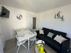 A living room with a dining table set for four, a black sofa with cushions, a TV mounted on the wall, and a decorative wall piece. - Charming apartment near beach - 4 guests (Cabourg)