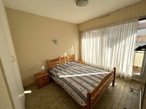 A bedroom with a wooden bed, a bedside table with a lamp, a wall-mounted light, and a window with sheer curtains. - Apartment sea and mountain view - 4 guests (Argelès-sur-Mer)