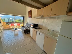 A compact kitchen with a dishwasher, a microwave, and a refrigerator. There is a dining area with a table and chairs, and a balcony with a view of buildings and trees. - Apartment near beach with parking - 5 guests (Argelès-sur-Mer)