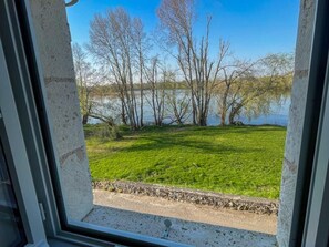 Une vue à travers une fenêtre sur un espace verdoyant, avec des arbres et une étendue d’eau.